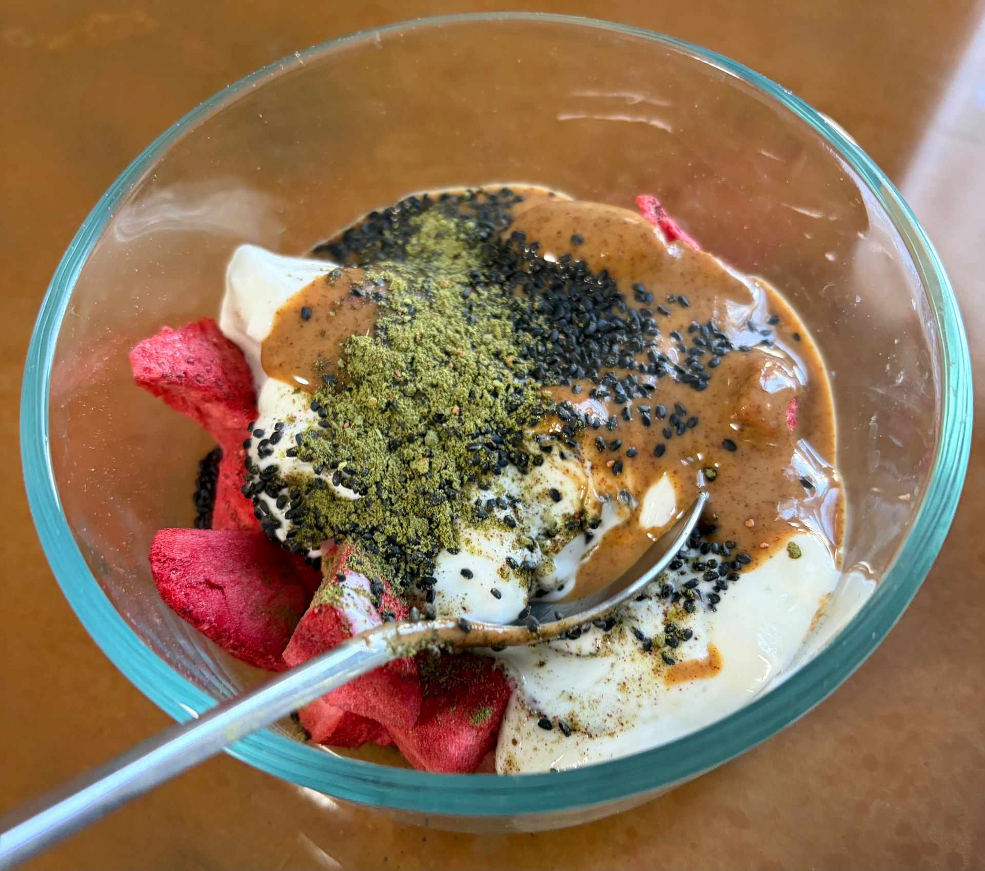 Glass bowl of Greek yogurt topped with berry greens powder, almond butter, basil seeds, and freeze-dried strawberries