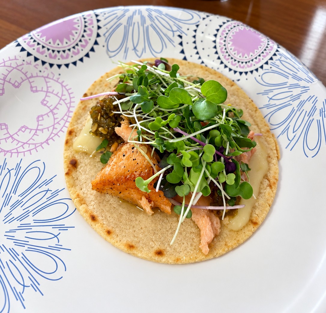 Gluten-free corn tortilla topped with seared trout, cheese, sweet jalapeño relish, and organic microgreens on a patterned paper plate